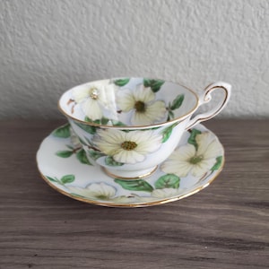 May include: A delicate teacup and saucer set. The white porcelain cup and saucer are adorned with a floral pattern featuring yellow and white flowers with green leaves. The rims are trimmed with gold.
