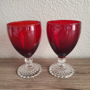 May include: Two matching goblets featuring deep red glass bowls and clear glass stems. The circular bases of the goblets are decorated with a raised hobnail pattern, providing a textured appearance to the bottom of each piece of glassware.