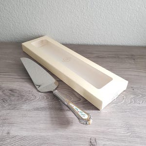May include: A silver cake server with a decorative handle featuring a gold trim and a holly design, next to a cream-colored box. The server's blade is shiny and reflects the light. The handle has red, green, and white accents.