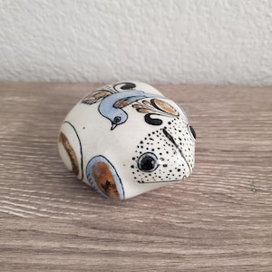 May include: A ceramic frog figurine with a white base, adorned with a blue bird and brown accents. The frog has black eyes and a dotted pattern on its face. The figurine is on a wooden surface.