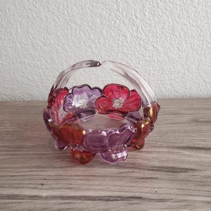 May include: A clear glass basket with a handle, adorned with red, pink, and purple floral designs. The basket has a scalloped edge and rests on three small feet. The flowers have gold accents.