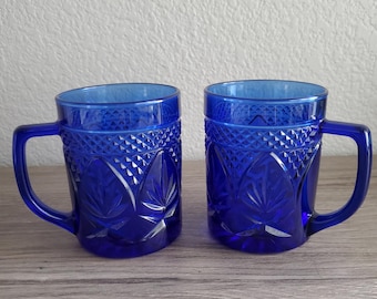 May include: Two cobalt blue glass mugs with handles. The mugs have a textured, cut-glass design featuring a repeating pattern of leaves and diamond shapes. The mugs are displayed on a wooden surface.