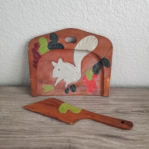 May include: A brown wooden wall-mounted holder featuring a carved white squirrel, green leaves, and red berries. A matching brown wooden serving utensil with a green leaf accent is positioned in front of the holder, which has a cutout handle.