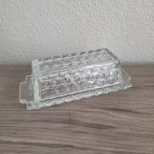 May include: A clear glass butter dish featuring a rectangular lid and base with a raised, geometric honeycomb pattern. The lid has a flat top and vertical sides, while the base includes small handles on each end for easy lifting and handling.