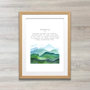 Op de afbeelding: Een ingelijst afdruk met een aquarel-illustratie van groene en blauwe bergen. De afdruk toont de definitie van het woord "Integrität" als "Moed kiezen boven comfort, kiezen wat juist is boven wat leuk, snel of gemakkelijk is; en onze waarden in de praktijk brengen in plaats van ze alleen te belijden."