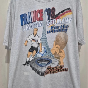 Puede incluir: Una camiseta gris claro de manga corta con un gr&aacute;fico de Francia '98 que muestra la Torre Eiffel, un jugador de f&uacute;tbol, un estadio, un trofeo y el texto "WM-TEAM GERMANY For the winning Soccer Adventure" en azul, negro, rojo y amarillo.