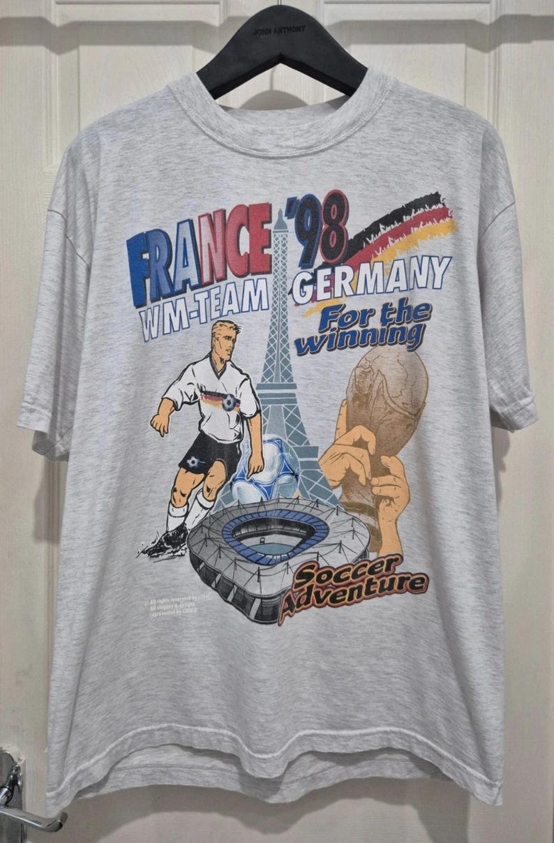 Puede incluir: Una camiseta gris claro de manga corta con un gr&aacute;fico de Francia '98 que muestra la Torre Eiffel, un jugador de f&uacute;tbol, un estadio, un trofeo y el texto "WM-TEAM GERMANY For the winning Soccer Adventure" en azul, negro, rojo y amarillo.