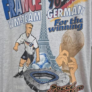 Puede incluir: Una camiseta gris claro con un estampado gr&aacute;fico con el texto France '98, WM-Team Germany, For the winning y Soccer Adventure. El dise&ntilde;o incluye una ilustraci&oacute;n de un jugador de f&uacute;tbol, un bal&oacute;n, la Torre Eiffel, un estadio y un trofeo.