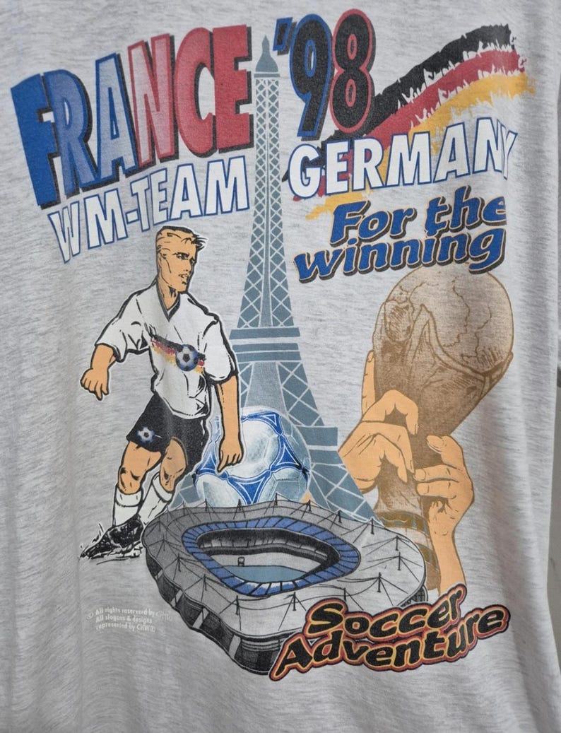 Puede incluir: Una camiseta gris claro con un estampado gr&aacute;fico con el texto France '98, WM-Team Germany, For the winning y Soccer Adventure. El dise&ntilde;o incluye una ilustraci&oacute;n de un jugador de f&uacute;tbol, un bal&oacute;n, la Torre Eiffel, un estadio y un trofeo.