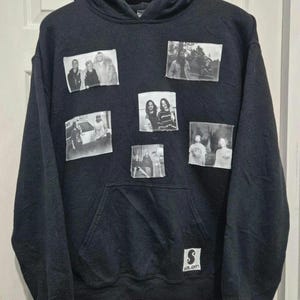 May include: Black hooded sweatshirt with a kangaroo pocket and a small white logo patch. The sweatshirt features several rectangular patches with black and white photographs. The hoodie is hanging on a black hanger.