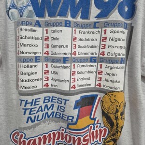 Puede incluir: Una camiseta gris claro con un gr&aacute;fico de la Copa Mundial de 1998. El dise&ntilde;o incluye un bal&oacute;n de f&uacute;tbol, el texto WM 98, una tabla que enumera los grupos de la A a la H con nombres de pa&iacute;ses en alem&aacute;n, el texto THE BEST TEAM IS NUMBER 1 y un trofeo.