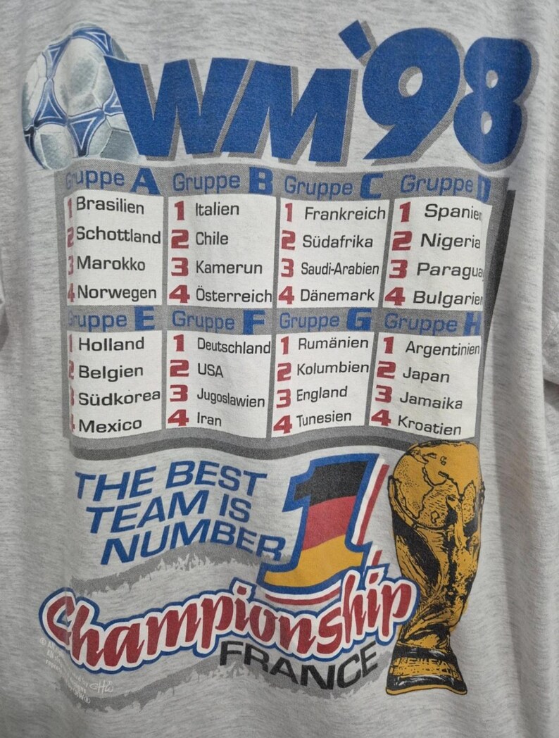 Puede incluir: Una camiseta gris claro con un gr&aacute;fico de la Copa Mundial de 1998. El dise&ntilde;o incluye un bal&oacute;n de f&uacute;tbol, el texto WM 98, una tabla que enumera los grupos de la A a la H con nombres de pa&iacute;ses en alem&aacute;n, el texto THE BEST TEAM IS NUMBER 1 y un trofeo.