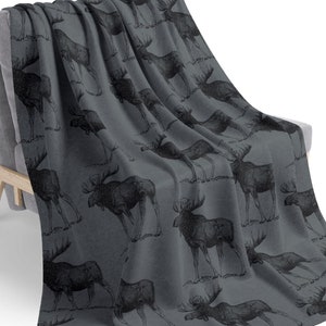 Puede incluir: Una manta de forro polar gris con un patrón repetido de siluetas de alces negras. La manta está drapeada sobre una silla gris.