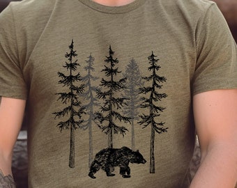 Può includere: T-shirt verde oliva con un'illustrazione nera di un orso che cammina davanti a una foresta di pini. Il design grafico è centrato sul petto. La maglietta è realizzata in un materiale morbido e confortevole.
