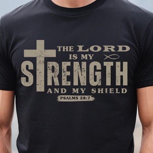 Puede incluir: Camiseta negra con un gráfico blanco desgastado de una cruz, las palabras "The Lord is my Strength and my Shield", y el texto "Psalms 28:7".