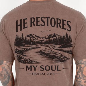 Könnte beinhalten: Braunes T-Shirt mit einem grafischen Design auf dem Rücken. Das Design zeigt eine Berglandschaft mit einem Fluss, Bäumen und dem Text "HE RESTORES MY SOUL - PSALM 23:3" in Schwarz.