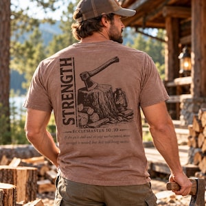 Può includere: Una t-shirt marrone a maniche corte con una grafica marrone scuro sul retro. La grafica raffigura un'ascia conficcata in un ceppo, una lanterna e legna accatastata. Il testo riporta STRENGTH, ECCLESIASTES 10:10 e un versetto su un'ascia.