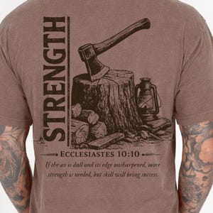 May include: Brown t-shirt with a graphic of an axe on a tree stump, a lantern, and the word "STRENGTH". The text "ECCLESIASTES 10:10" and a quote about strength are also printed on the shirt.