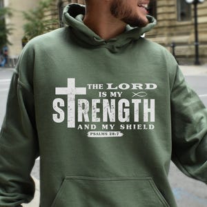 Puede incluir: Una sudadera con capucha verde con un estampado gráfico blanco que dice "The Lord is my Strength and my Shield, Psalms 28:7".
