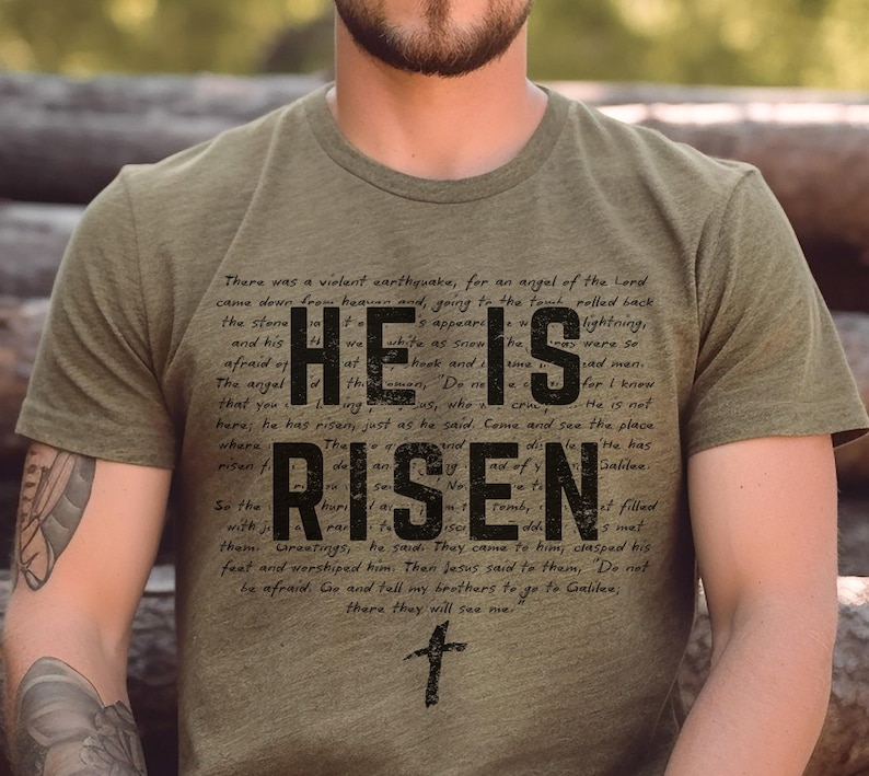 K&ouml;nnte beinhalten: Olivgr&uuml;nes T-Shirt mit dem Schriftzug "HE IS RISEN" in gro&szlig;en, schwarzen Buchstaben. Der Text ist von religi&ouml;sem Text und einem kleinen Kreuz umgeben.