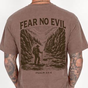 以下が含まれることがあります： 背面全体に「FEAR NO EVIL」の文字がプリントされた茶色のTシャツ。その下には、渓谷を歩くハイカーの詳細なイラストが描かれています。「PSALM 23:4」の文字がイラストの下に印刷されています。