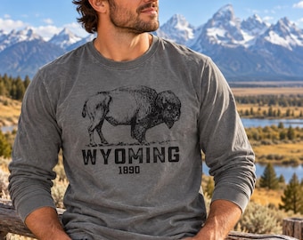 May include: A charcoal gray long-sleeve shirt featuring a black graphic of a bison in profile. Below the bison, the text reads WYOMING in a bold, block font, followed by the year 1890 in a smaller, matching font centered on the chest.