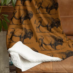 May include: A brown and white fleece throw blanket with a repeating pattern of black buffalo silhouettes. The blanket is folded and draped over a brown leather couch.