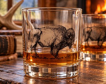 May include: A clear glass tumbler featuring a black etched illustration of a bison in profile. The glass contains an amber-colored liquid. A second identical glass with a bison illustration is partially visible in the background.