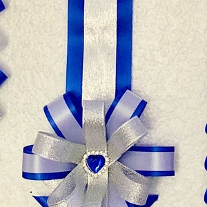 Puede incluir: Una larga decoraci&oacute;n de cinta vertical con el texto Diamond Braid. Consta de una franja central plateada flanqueada por cintas azul real, que termina en un patr&oacute;n de chevr&oacute;n en capas y un lazo central con una gema azul en forma de coraz&oacute;n.