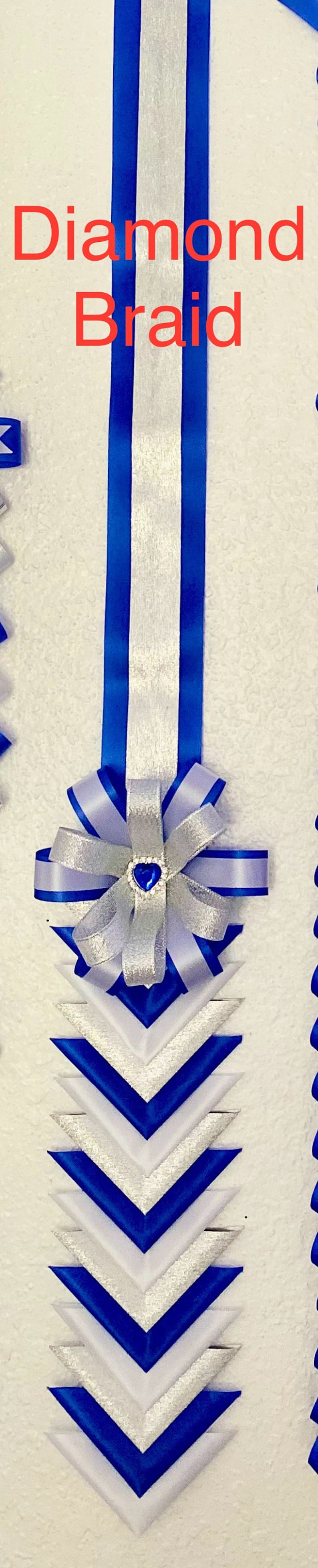 Puede incluir: Una larga decoraci&oacute;n de cinta vertical con el texto Diamond Braid. Consta de una franja central plateada flanqueada por cintas azul real, que termina en un patr&oacute;n de chevr&oacute;n en capas y un lazo central con una gema azul en forma de coraz&oacute;n.