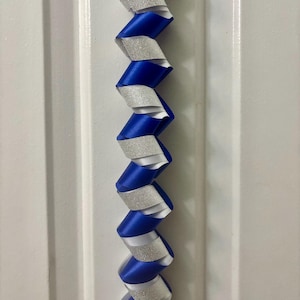 Puede incluir: Una guirnalda decorativa vertical con un patr&oacute;n de espiral repetitivo que alterna cinta de sat&eacute;n azul real y cinta de purpurina plateada. Las cintas est&aacute;n trenzadas para crear una estructura continua en forma de sacacorchos.