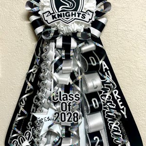 Puede incluir: Un adorno de homecoming mum en negro, blanco y plata con un emblema central en forma de escudo con la letra S y el texto KNIGHTS. El dise&ntilde;o incluye cintas en cascada, un recorte de estrella con el texto Class Of 2028 y letras adicionales.