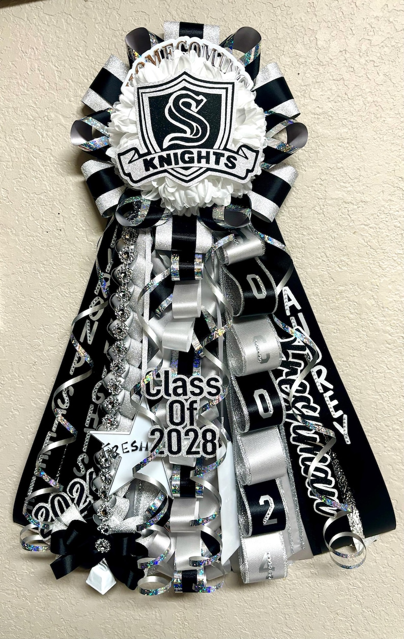 Puede incluir: Un adorno de homecoming mum en negro, blanco y plata con un emblema central en forma de escudo con la letra S y el texto KNIGHTS. El dise&ntilde;o incluye cintas en cascada, un recorte de estrella con el texto Class Of 2028 y letras adicionales.