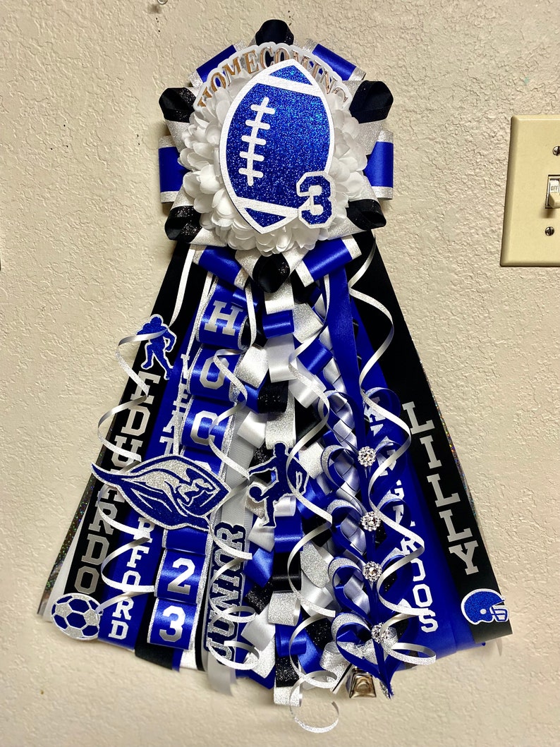 Puede incluir: Un adorno de homecoming mum en azul, negro y blanco con un recorte de bal&oacute;n de f&uacute;tbol americano brillante con el n&uacute;mero 3, cintas en capas y varios detalles deportivos que incluyen siluetas de atletas, un bal&oacute;n de f&uacute;tbol y los nombres LILLY y GUNNER.