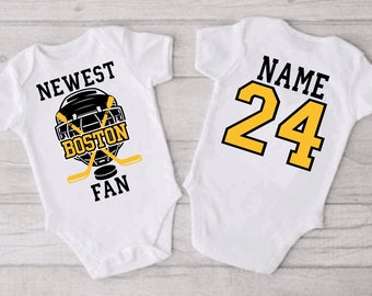 May include: White baby onesie with a black and yellow hockey design. The onesie says "Newest Boston Fan" and has a hockey helmet, sticks, and puck. The back of the onesie has a yellow number 24 and the word "Name".