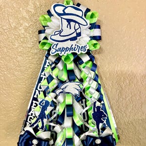 Puede incluir: Un adorno de homecoming mum con capas de cintas de color verde lima, azul marino y plateado. Incluye un recorte de sombrero de vaquero azul brillante con el texto Sapphires, un emblema de &aacute;guila y dos siluetas de bailarines, todo suspendido de un mosquet&oacute;n negro.