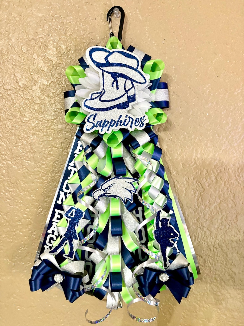 Puede incluir: Un adorno de homecoming mum con capas de cintas de color verde lima, azul marino y plateado. Incluye un recorte de sombrero de vaquero azul brillante con el texto Sapphires, un emblema de &aacute;guila y dos siluetas de bailarines, todo suspendido de un mosquet&oacute;n negro.