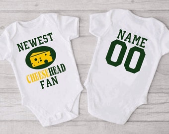 May include: White baby onesie with green and yellow text and a cheese graphic. The text reads "NEWEST CHEESEHEAD FAN". The back of the onesie has a green number "00" and the text "NAME".
