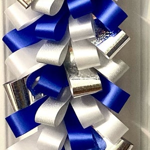 Puede incluir: Una decoraci&oacute;n de trenza de bucles vertical construida con bucles alternos de cinta azul real s&oacute;lida, cinta blanca s&oacute;lida y cinta hologr&aacute;fica plateada. Las cintas est&aacute;n superpuestas y dobladas para crear un patr&oacute;n denso, texturizado y repetitivo.