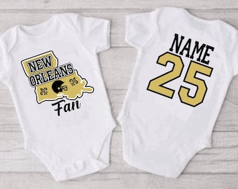 May include: Two white baby onesies. One onesie has a gold outline of the state of Louisiana with the text "New Orleans Fan" and a football helmet graphic. The other onesie has the text "NAME 25" in gold with a black outline.