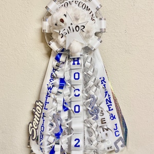 Puede incluir: Un adorno de homecoming mum con un oso de peluche blanco, una estrella recortada con la etiqueta SENIOR y cintas en cascada en blanco, azul y plata. Las cintas muestran textos como HOCO 2023, RIANE & JC y SENIOR, con dijes de notas musicales azules.