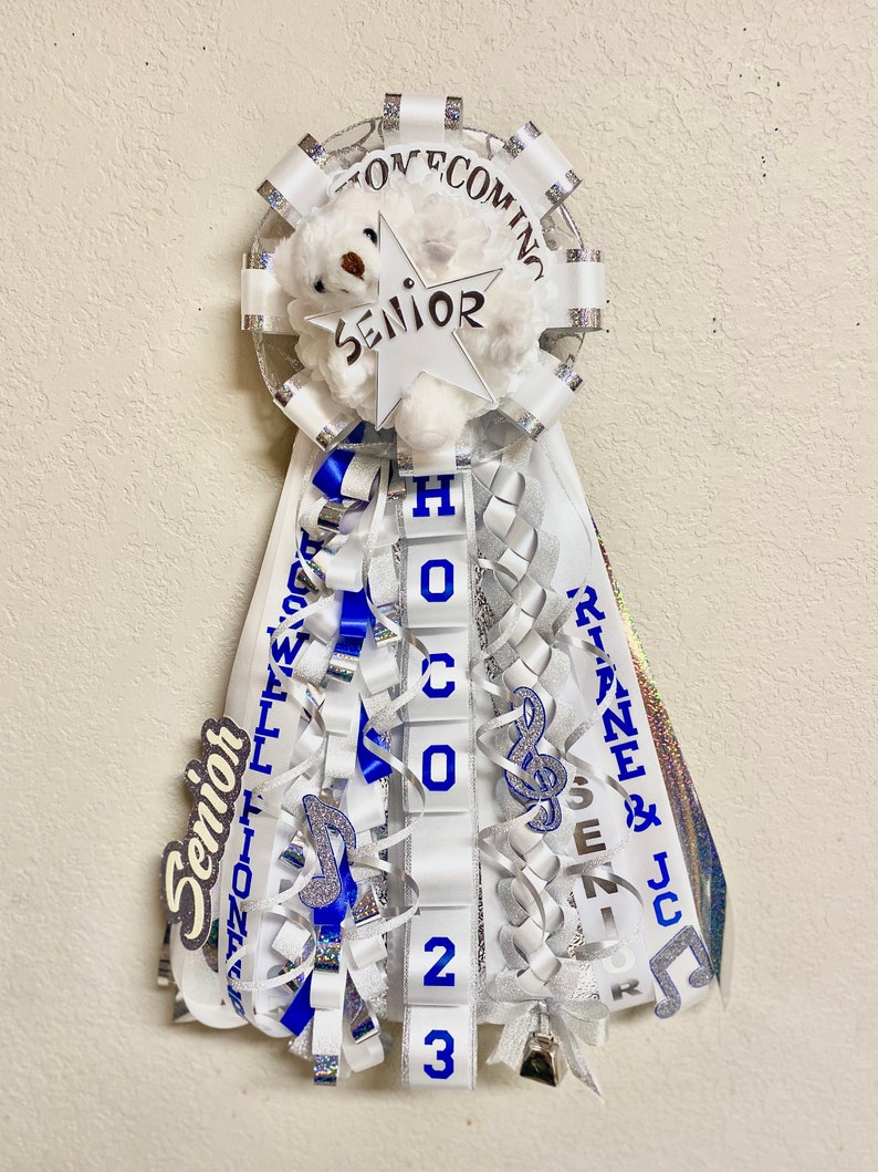 Puede incluir: Un adorno de homecoming mum con un oso de peluche blanco, una estrella recortada con la etiqueta SENIOR y cintas en cascada en blanco, azul y plata. Las cintas muestran textos como HOCO 2023, RIANE & JC y SENIOR, con dijes de notas musicales azules.