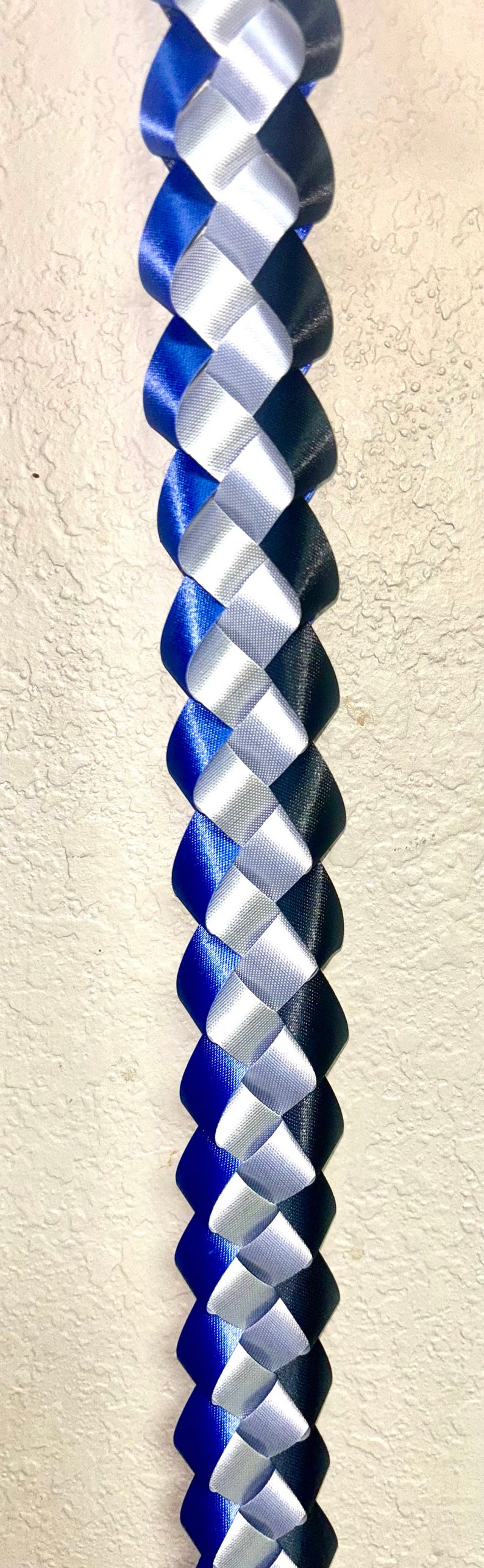 Puede incluir: Una trenza de cinta decorativa larga que presenta un patr&oacute;n repetitivo de cintas de sat&eacute;n azul real, plateado y negro tejidas juntas en un dise&ntilde;o en espiral apretado y entrelazado que crea una hebra vertical continua, texturizada y multicolor.