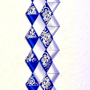 Puede incluir: Una larga decoraci&oacute;n de trenza de victoria vertical hecha de cintas entrelazadas de color azul real y plata. Las cintas azules presentan un patr&oacute;n de filigrana blanca en espiral. La parte inferior termina en una roseta de cinta.