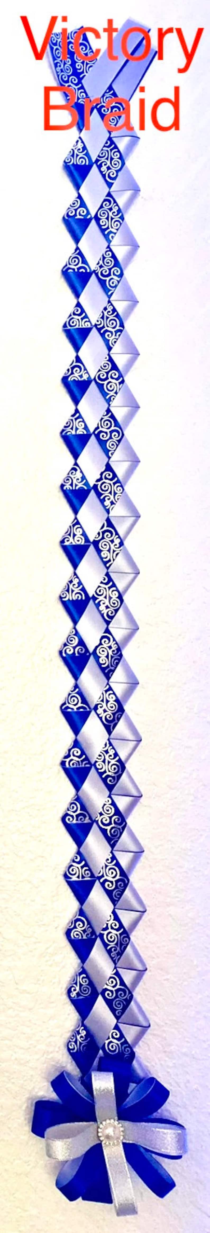 Puede incluir: Una larga decoraci&oacute;n de trenza de victoria vertical hecha de cintas entrelazadas de color azul real y plata. Las cintas azules presentan un patr&oacute;n de filigrana blanca en espiral. La parte inferior termina en una roseta de cinta.