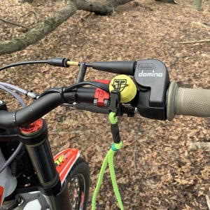 May include: Close-up of a black motorcycle handlebar with a Domino throttle, a red lever, and a yellow throttle lock. The handlebar is attached to a white motorcycle.