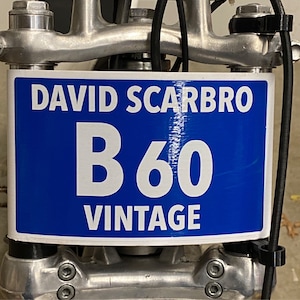 May include: A blue and white sign with the text "DAVID SCARBRO B60 VINTAGE" is mounted on a silver metal structure. The sign is rectangular with white text on a blue background. The metal structure has bolts and other mechanical components.