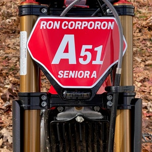 May include: Close-up of a red and black motorcycle. The front features a red plate with "RON CORPORON", "A51", and "SENIOR A" in white. The handlebars are black, and the suspension forks are gold.