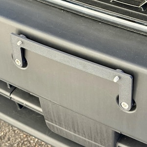 Puede incluir: Un soporte negro para la placa de matrícula está montado en el parachoques de un vehículo. El soporte rectangular tiene esquinas redondeadas y se fija con tornillos plateados. El parachoques es negro y tiene una superficie texturizada.