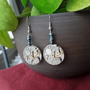 May include: A pair of silver earrings with teal blue beads and circular watch mechanisms. The watch mechanisms are made of silver and gold metal and have visible gears.